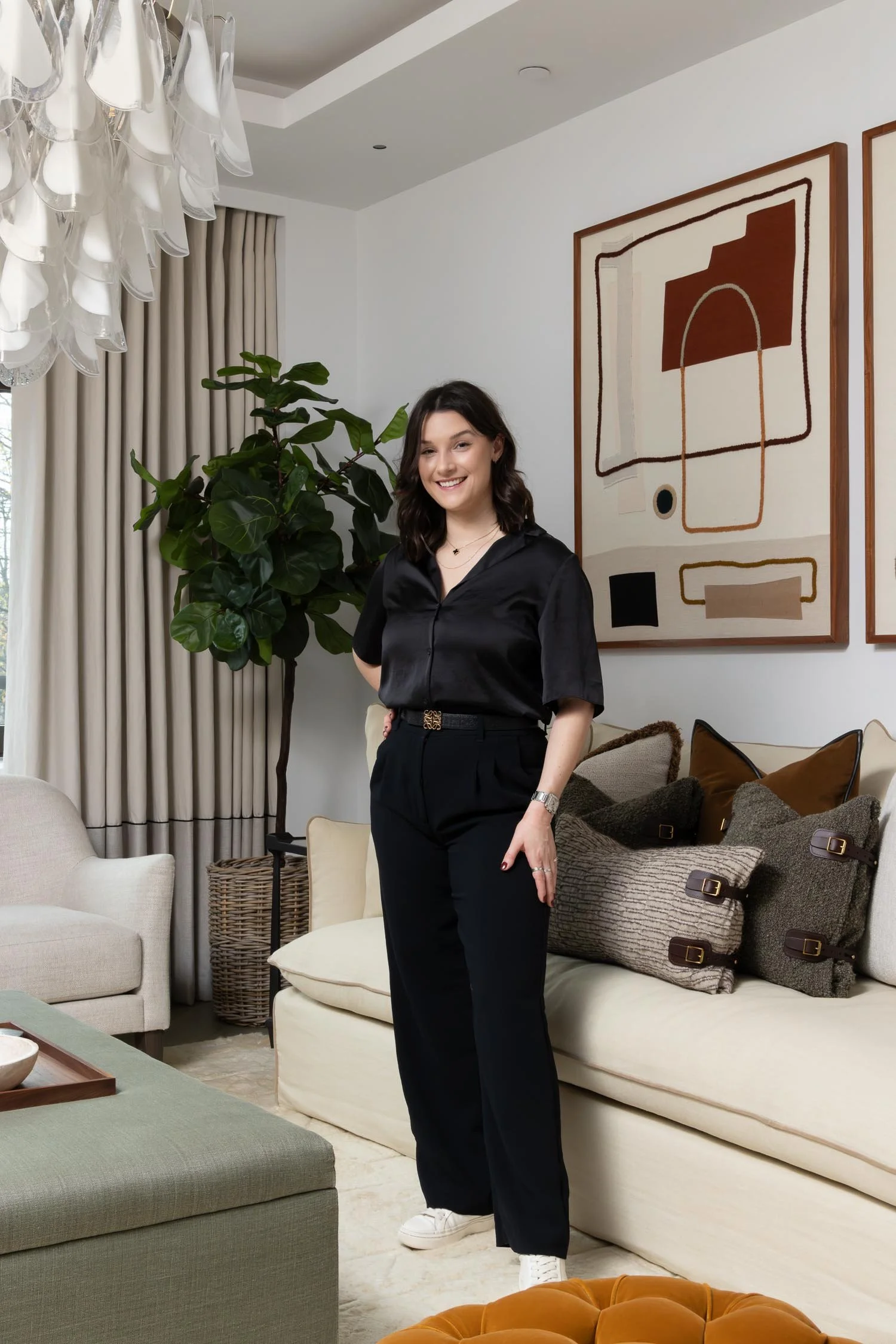 Woman in a black silk shirt and trousers smiles while standing in a modern living room with abstract art and a large plant