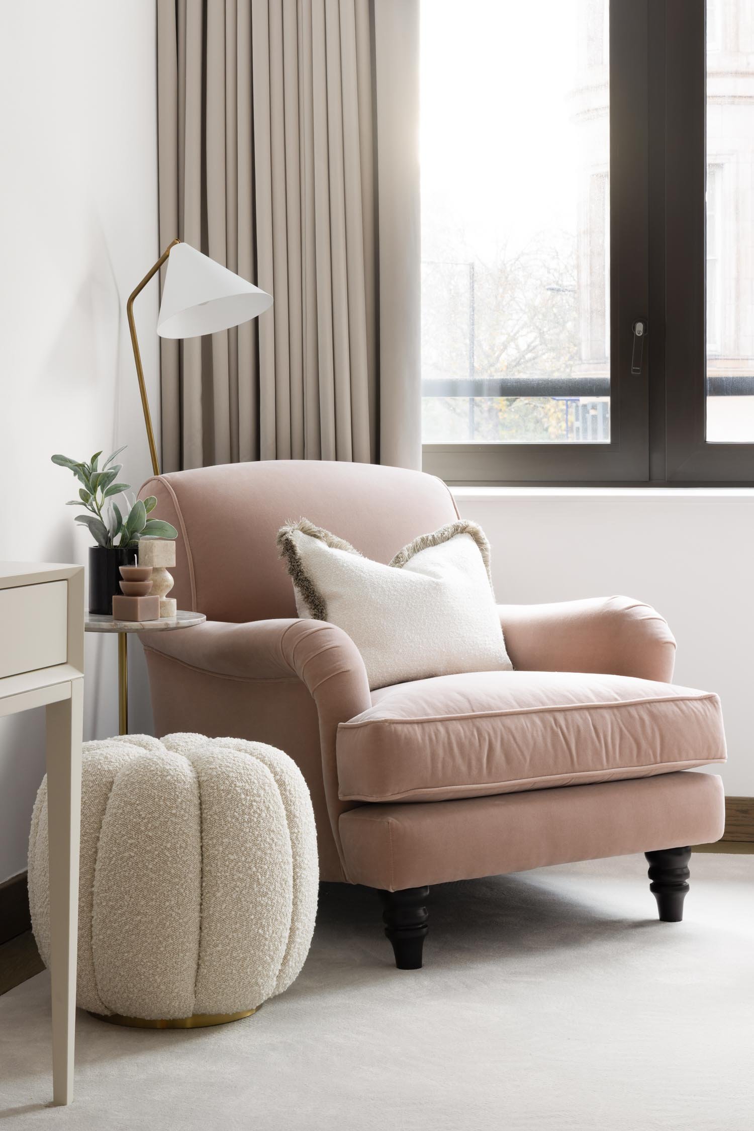 Dusty pink velvet armchair with a white textured cushion and footstool next to a large window with neutral curtains