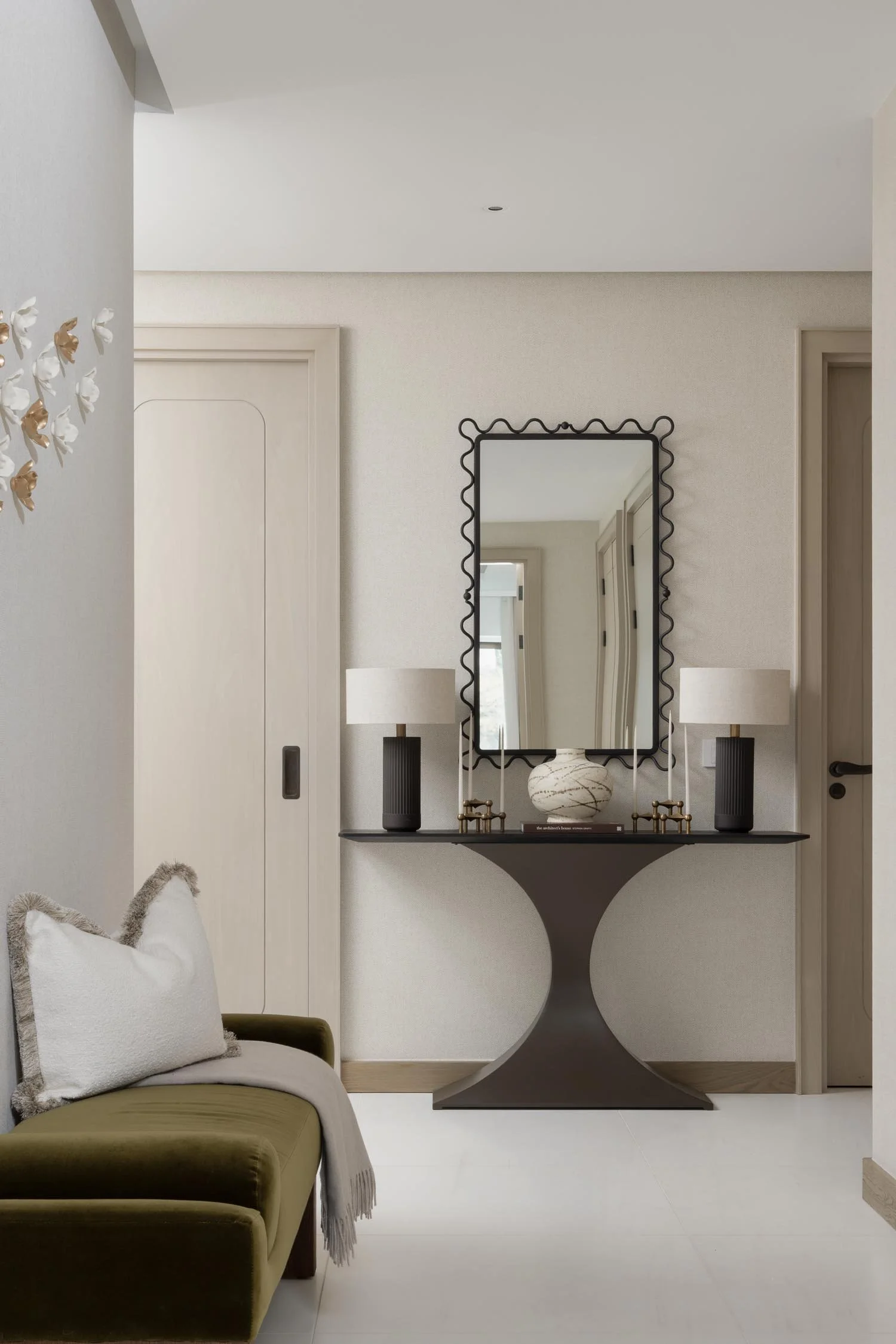 Dark hallway console table with a wavy-edged mirror and two black lamps against a neutral textured wallpaper background