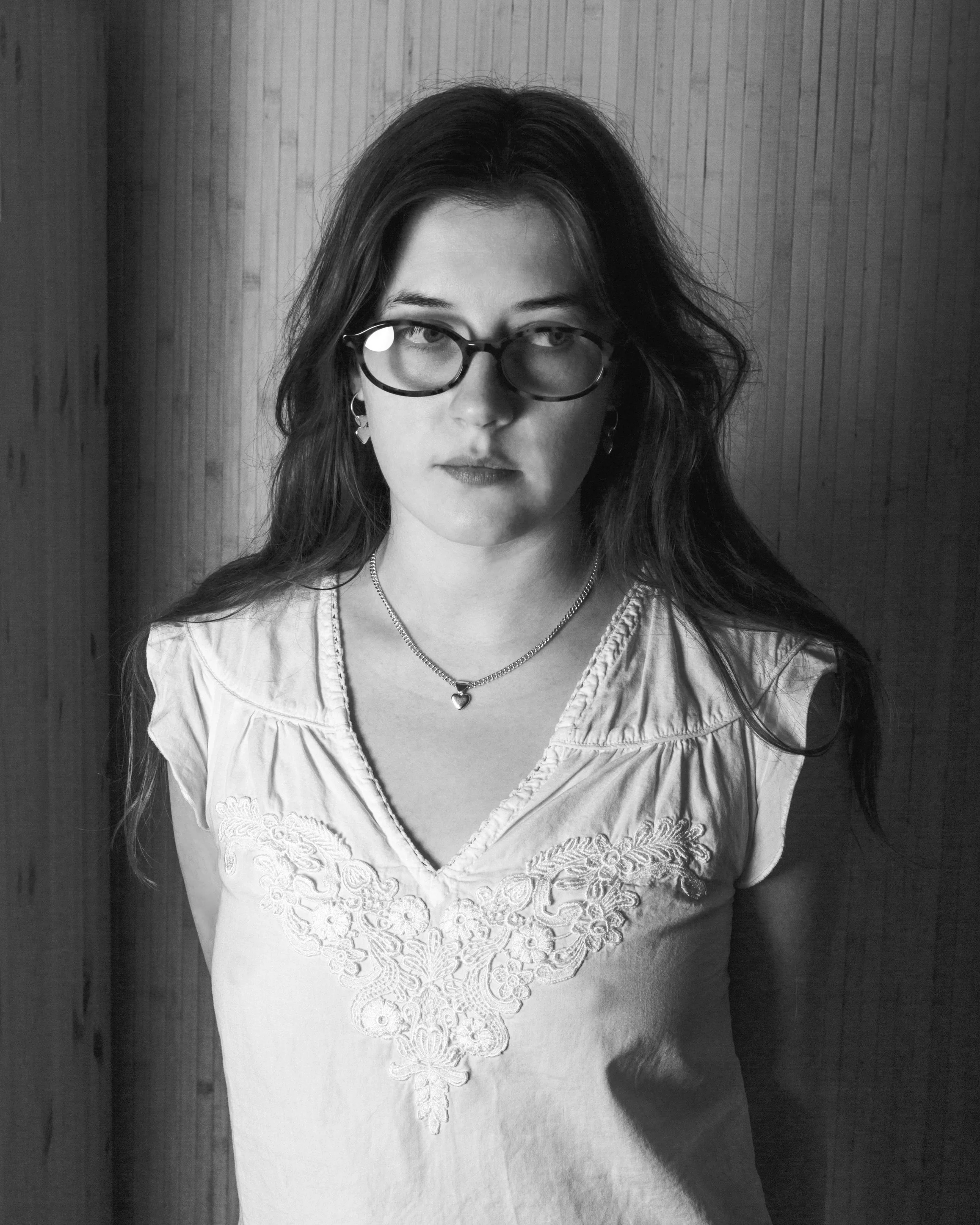 Black and white photo of a young woman with long dark hair, wearing glasses, earrings, a necklace with a heart pendant, and a embroidered top, standing against a wooden wall.