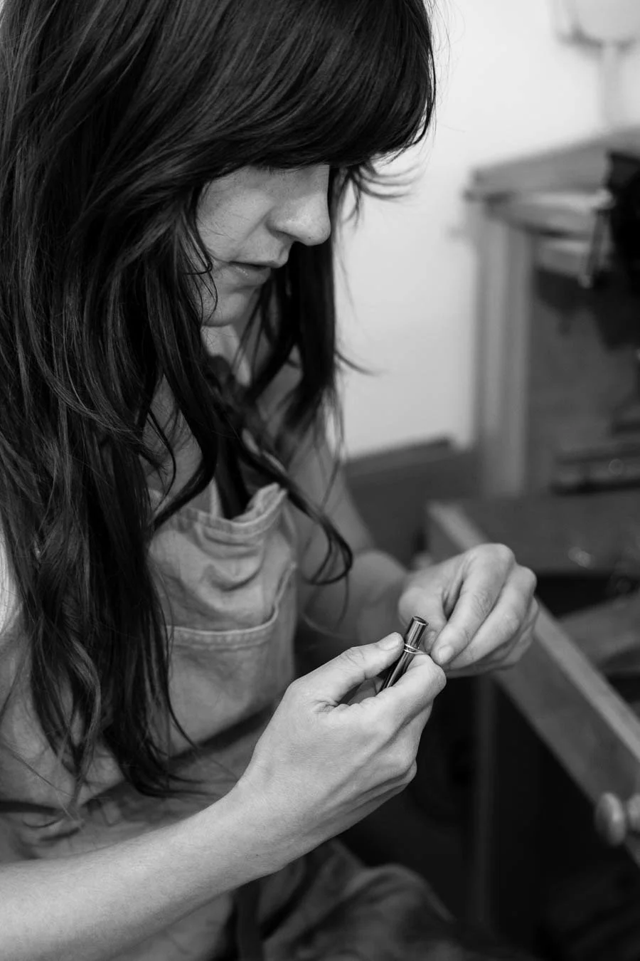 A woman with long dark hair, wearing an apron, working on a metal object with a small tool in a workshop.
