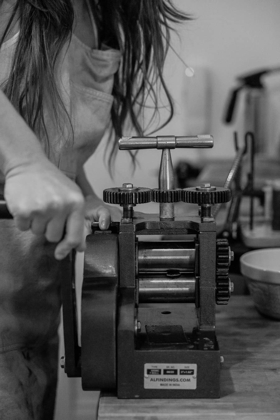 Black and white photo of a person with long hair operating a small vintage mechanical machine with gears and rollers. The person is wearing a light-colored shirt, and the machine is placed on a wooden surface.