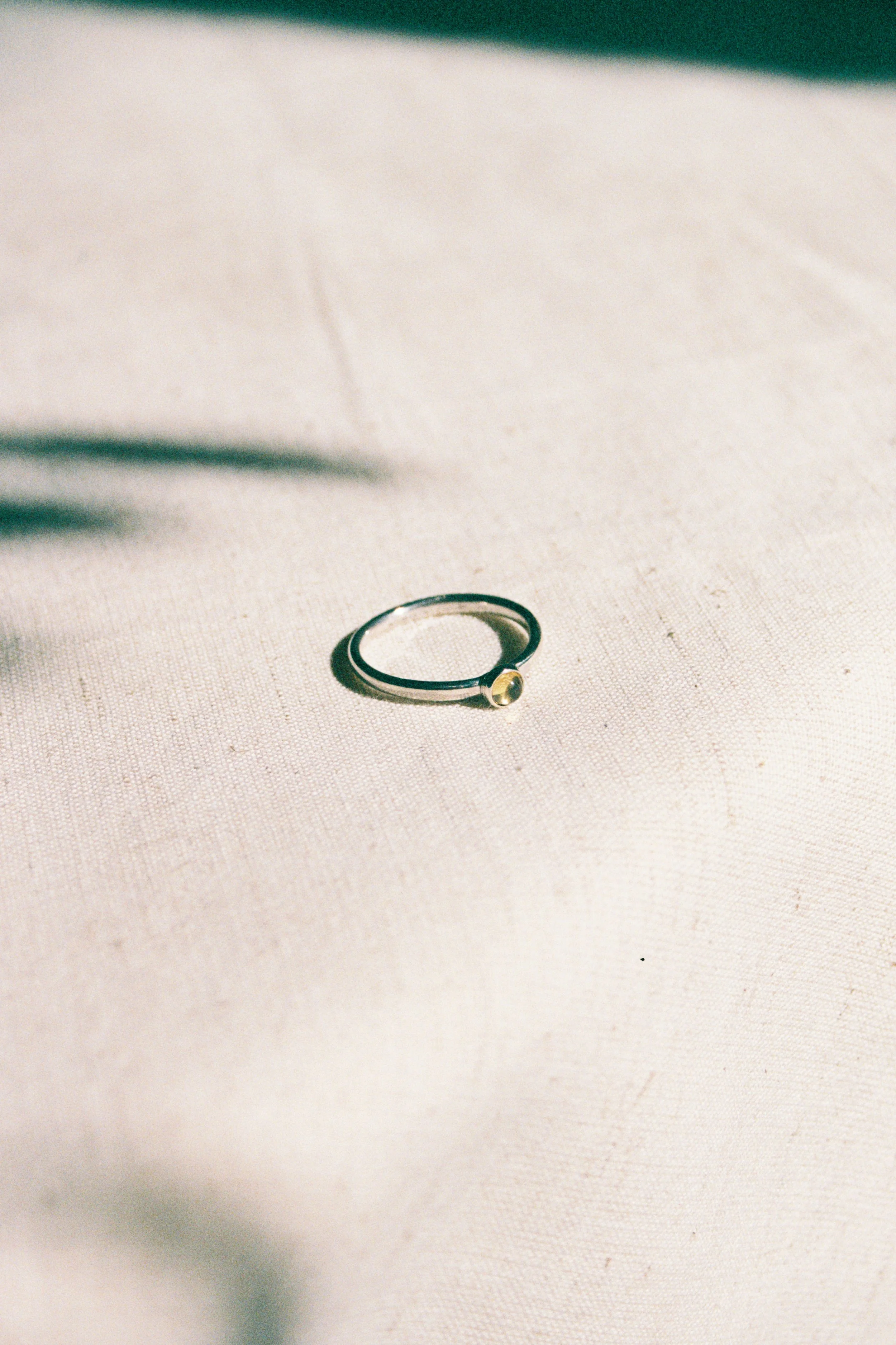 A ring with a silver band and a small yellow gemstone resting on a beige fabric surface, with a shadow and blurred plant leaves in the background.