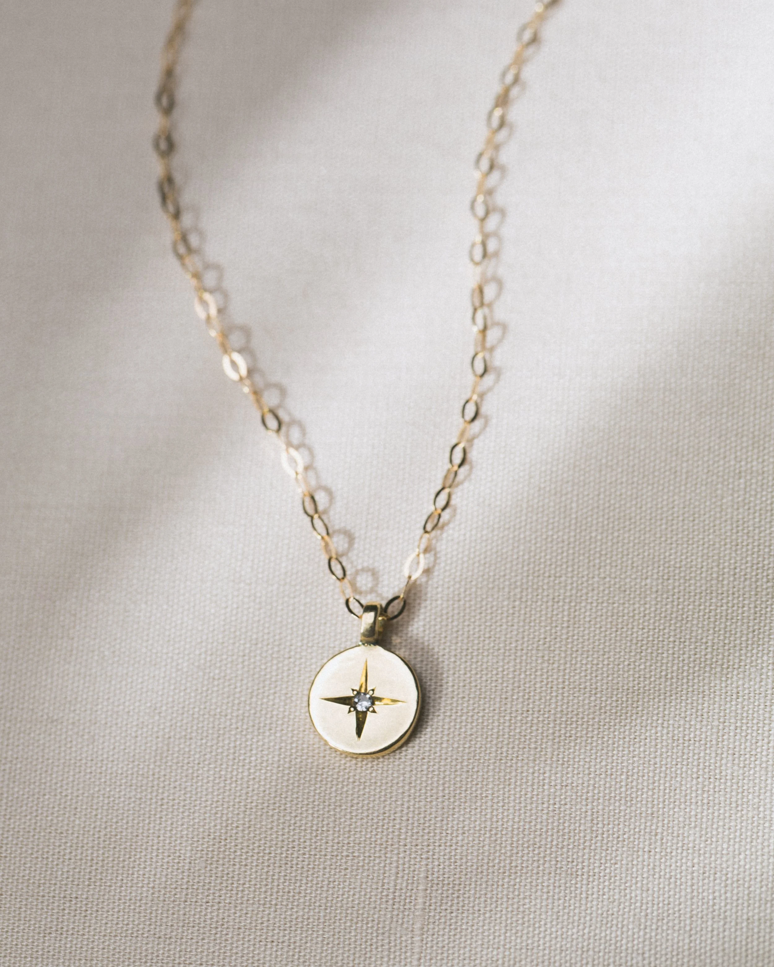 Gold necklace with a round pendant displaying a compass design with a small gemstone in the center, resting on a light-colored fabric background.