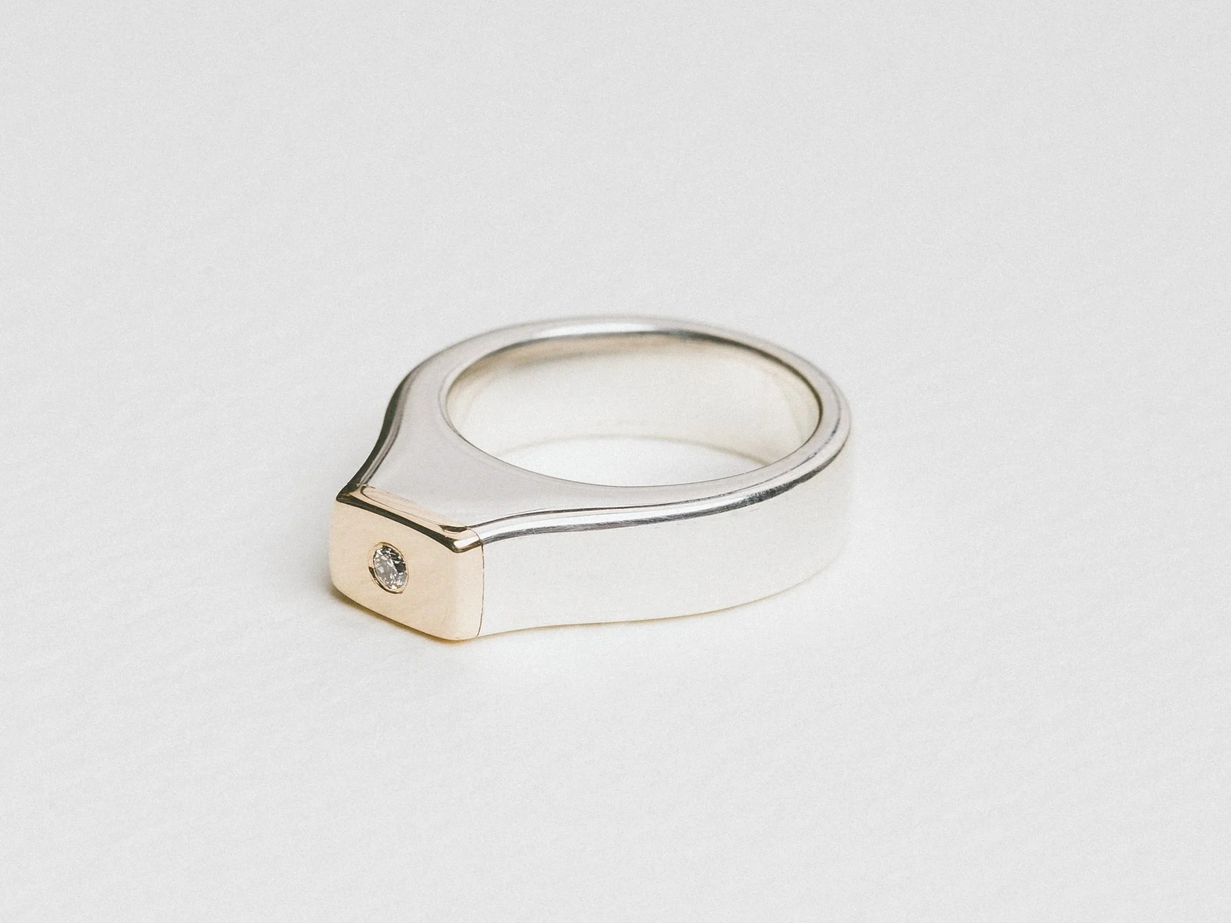 A silver and gold ring with a small diamond inset on the front, placed on a plain white background.