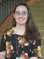 A woman with long brown hair and glasses wearing a floral dress, standing in front of stairs.