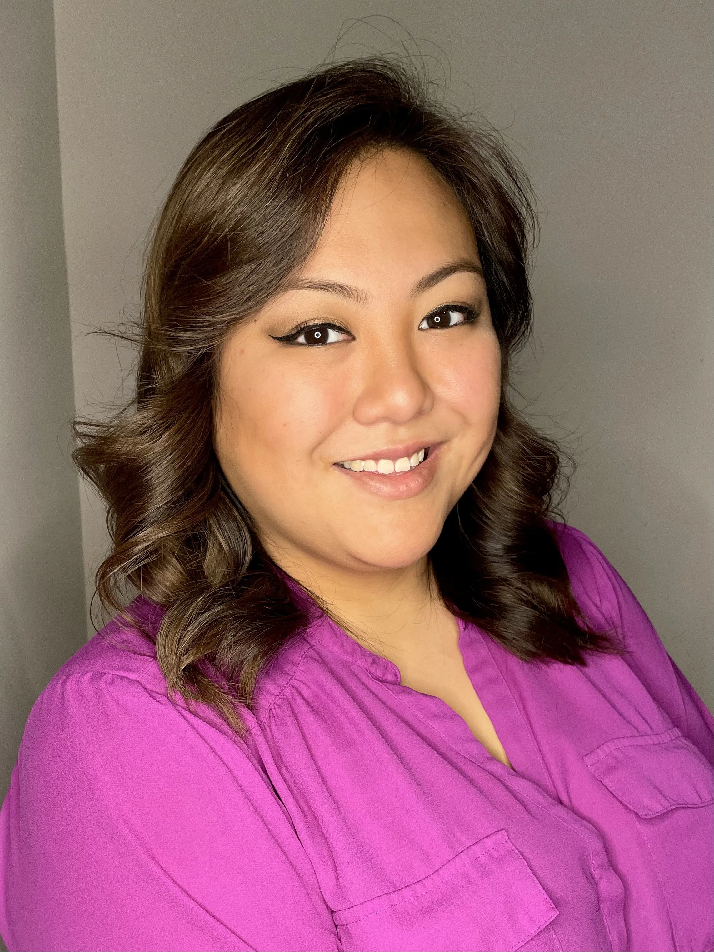 Portrait of a person with long, wavy hair wearing a purple blouse, smiling against a gray background.