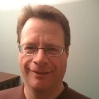 A person wearing glasses and a brown shirt smiling at the camera against a light-colored wall.