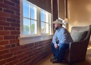 Man in cowboy hat sitting in a chair by a window with brick wall, looking outside, relaxed setting.