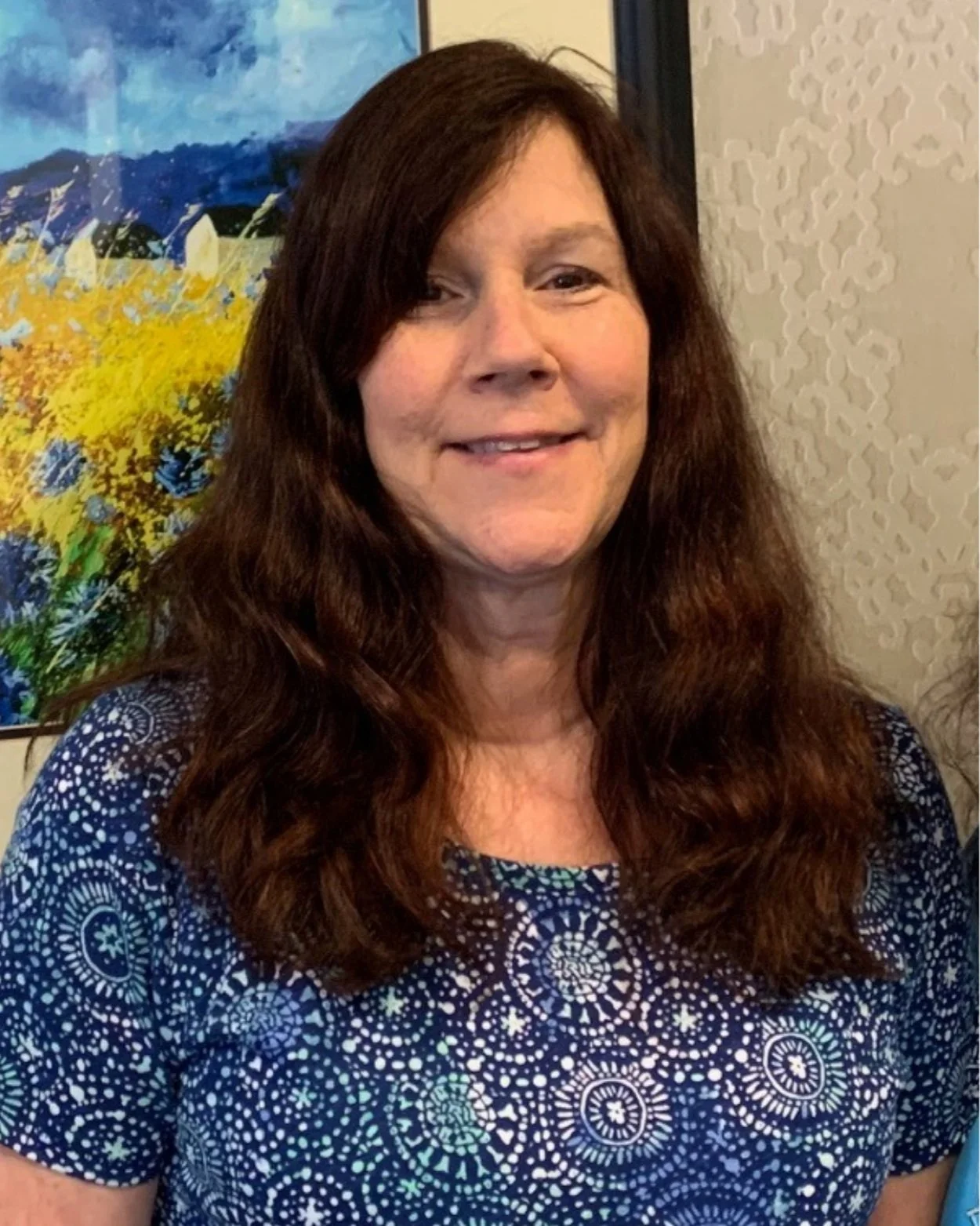 Smiling woman with long curly brown hair, wearing a patterned blue shirt, standing indoors with a colorful painting in the background.