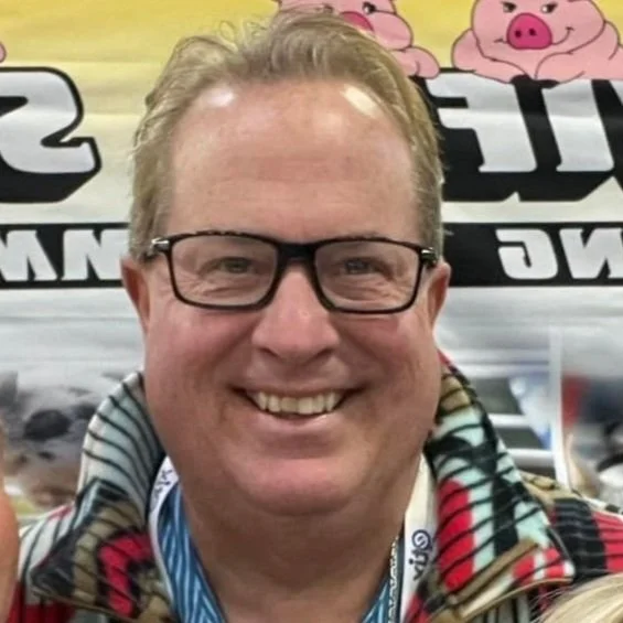 A smiling man wearing glasses and a colorful plaid jacket against a background with cartoon pigs and partial signage.
