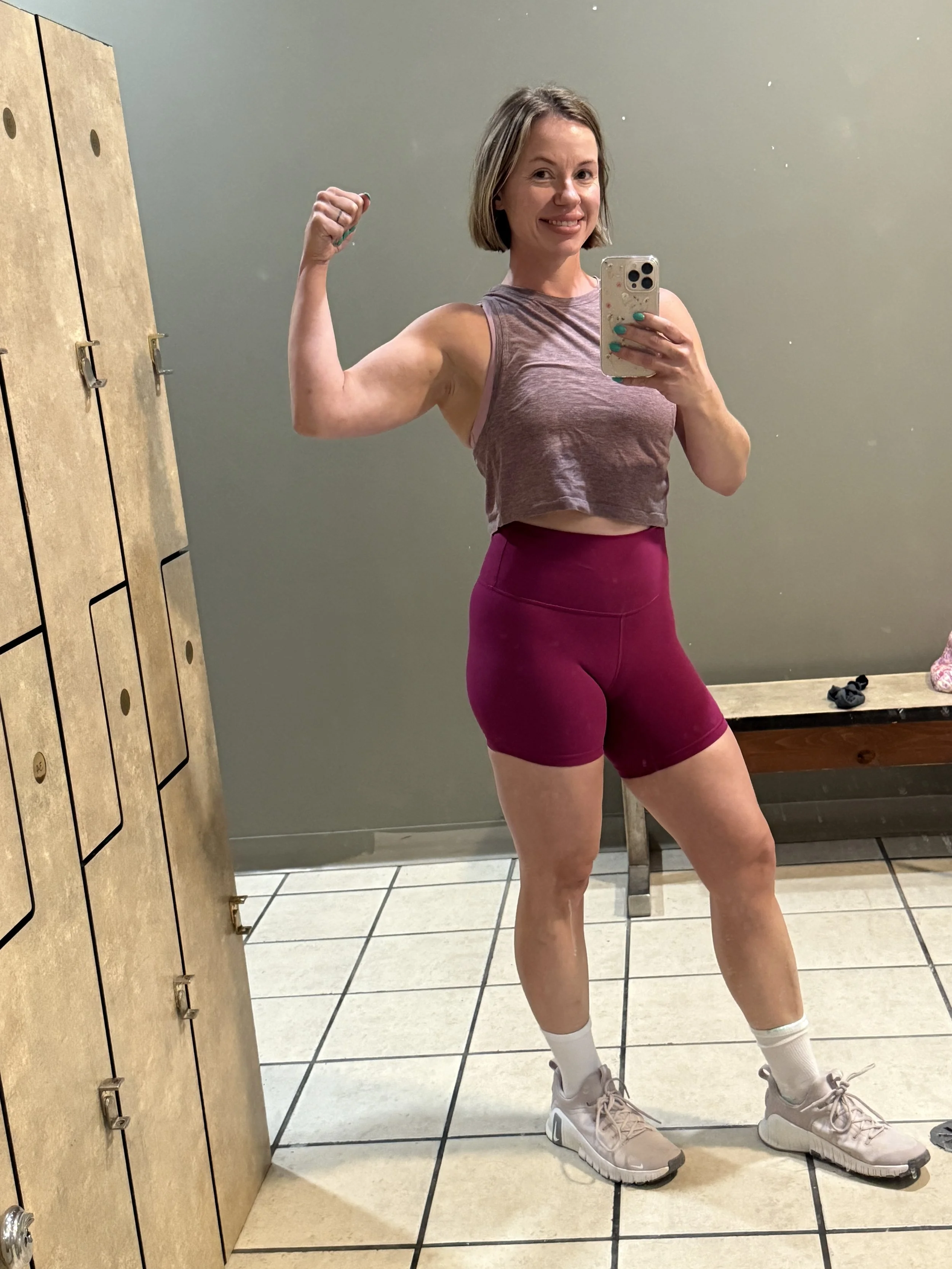 A woman taking a selfie in workout gear, flexing her arm muscles, and smiling.