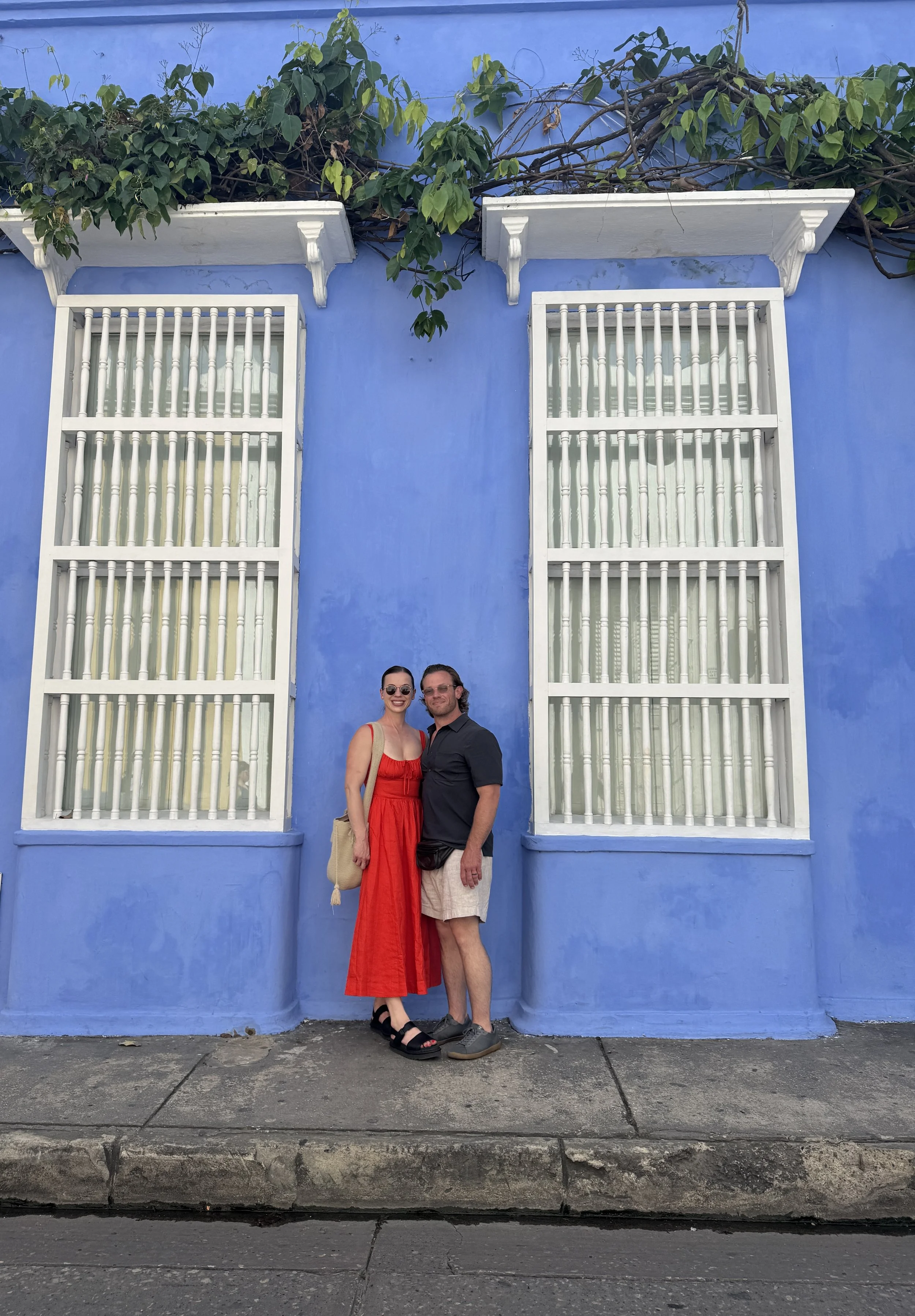 A couple stands in front of a vibrant blue building with white-framed windows and decorative overhangs, surrounded by green foliage.