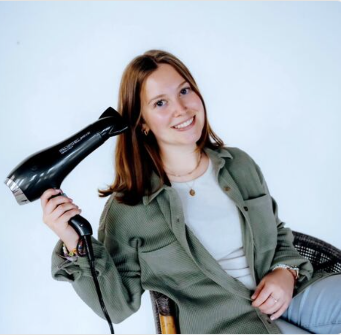 A woman with shoulder-length red hair smiling while holding a black hairdryer near her head, sitting on a chair against a light background.