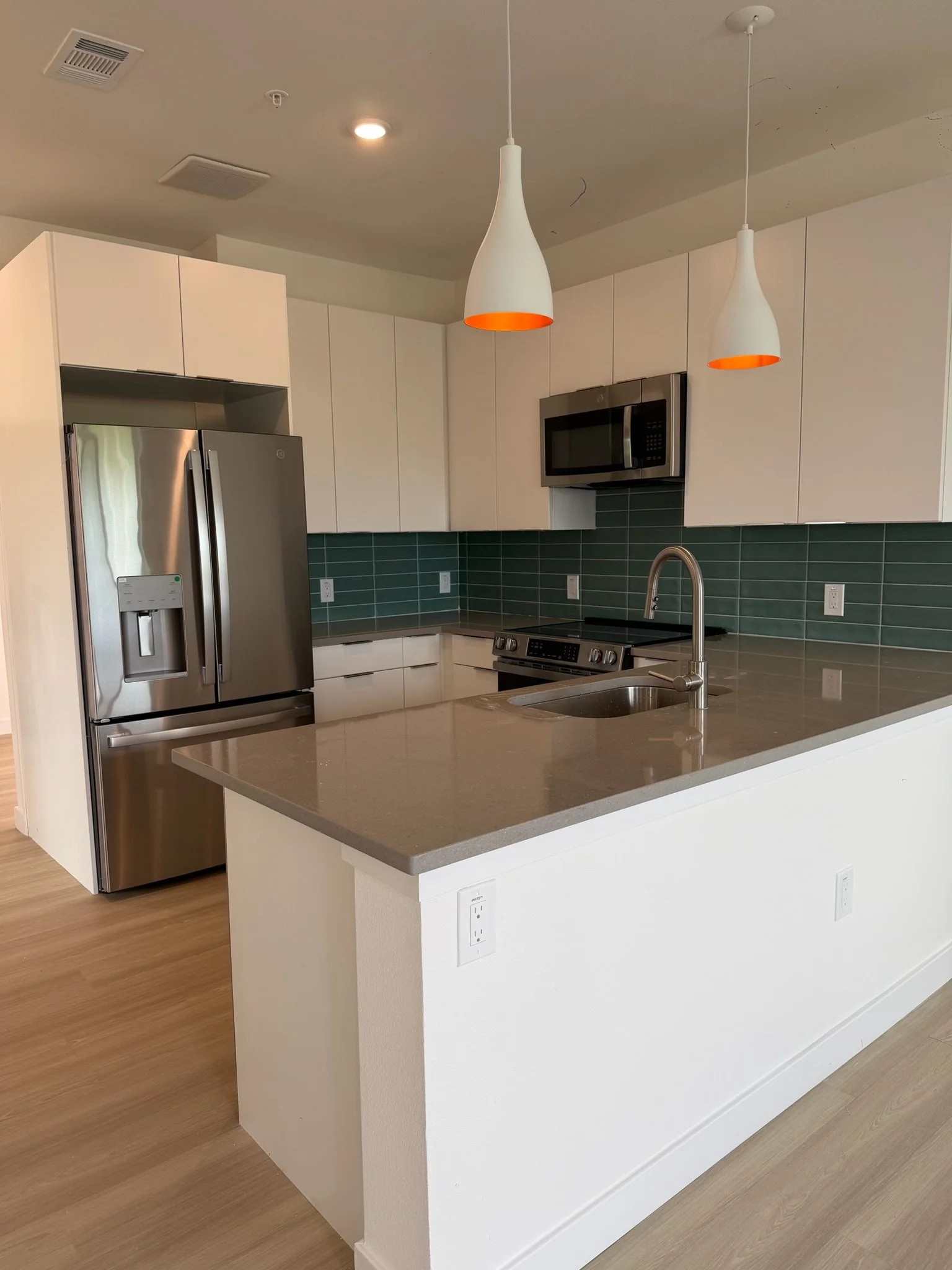 Modern kitchen with white cabinets, stainless steel refrigerator, microwave, green tiled backsplash, kitchen sink, and pendant lights with hardwood floors. 