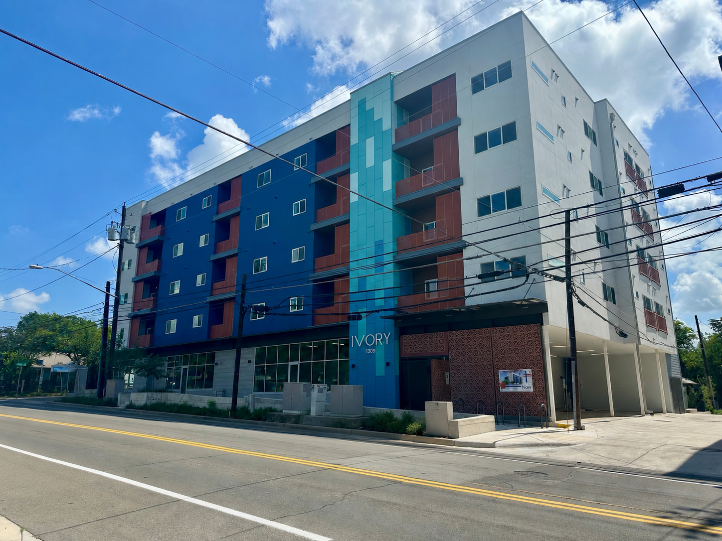 A modern multi-story residential building with a blue and white facade, featuring multiple balconies, large windows, and a glass entrance area labeled 'IVORY' with the number 1309. The building is located on a street with clear blue sky. 