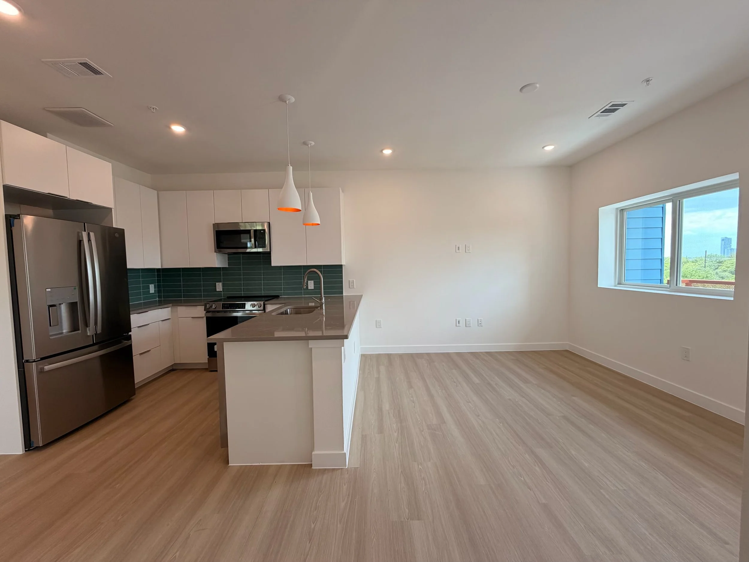 A modern kitchen with white cabinets, stainless steel appliances, a green tiled backsplash, light wood flooring, a kitchen island, and a large window bringing in natural light.