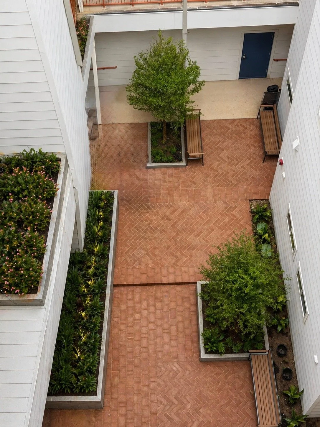 Top-down view of a small courtyard with brick paving, benches, trees, and surrounding white buildings with windows and plants.