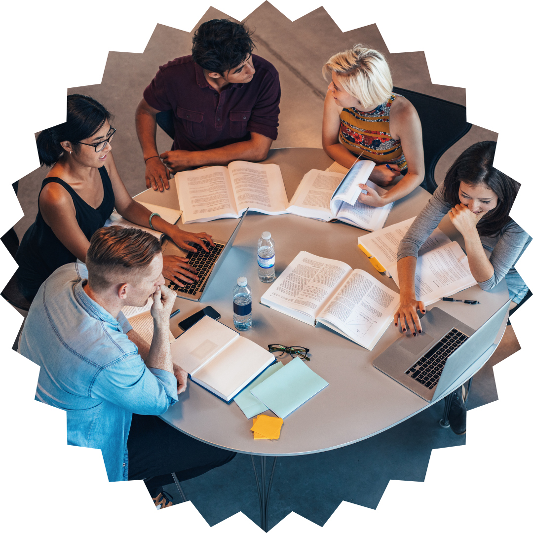 Five students studying together around a round table covered in open books, laptops and notes, viewed from above.