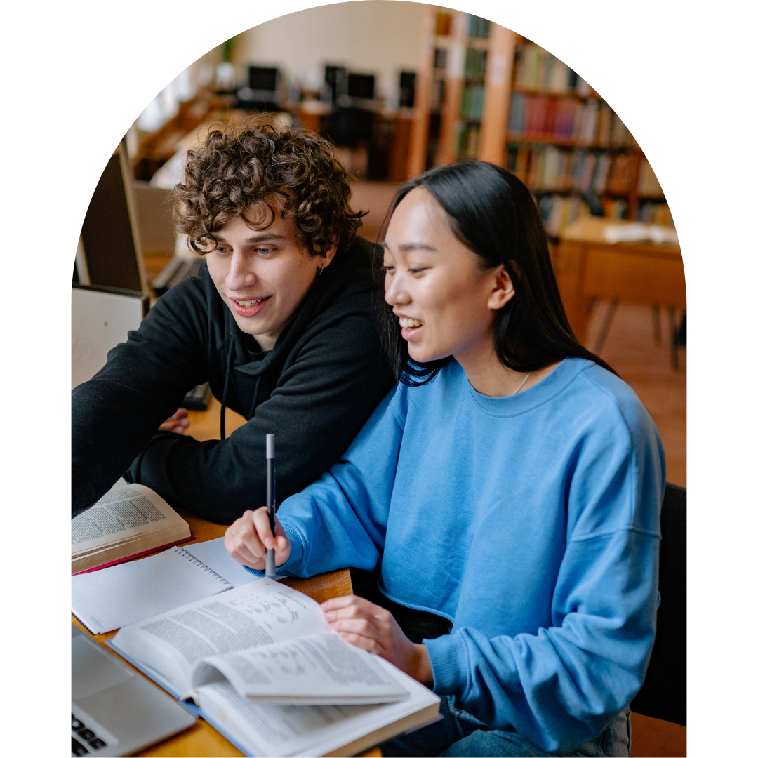 Two students sitting side by side in a library, smiling and looking at an open textbook together, with bookshelves visible in the background.