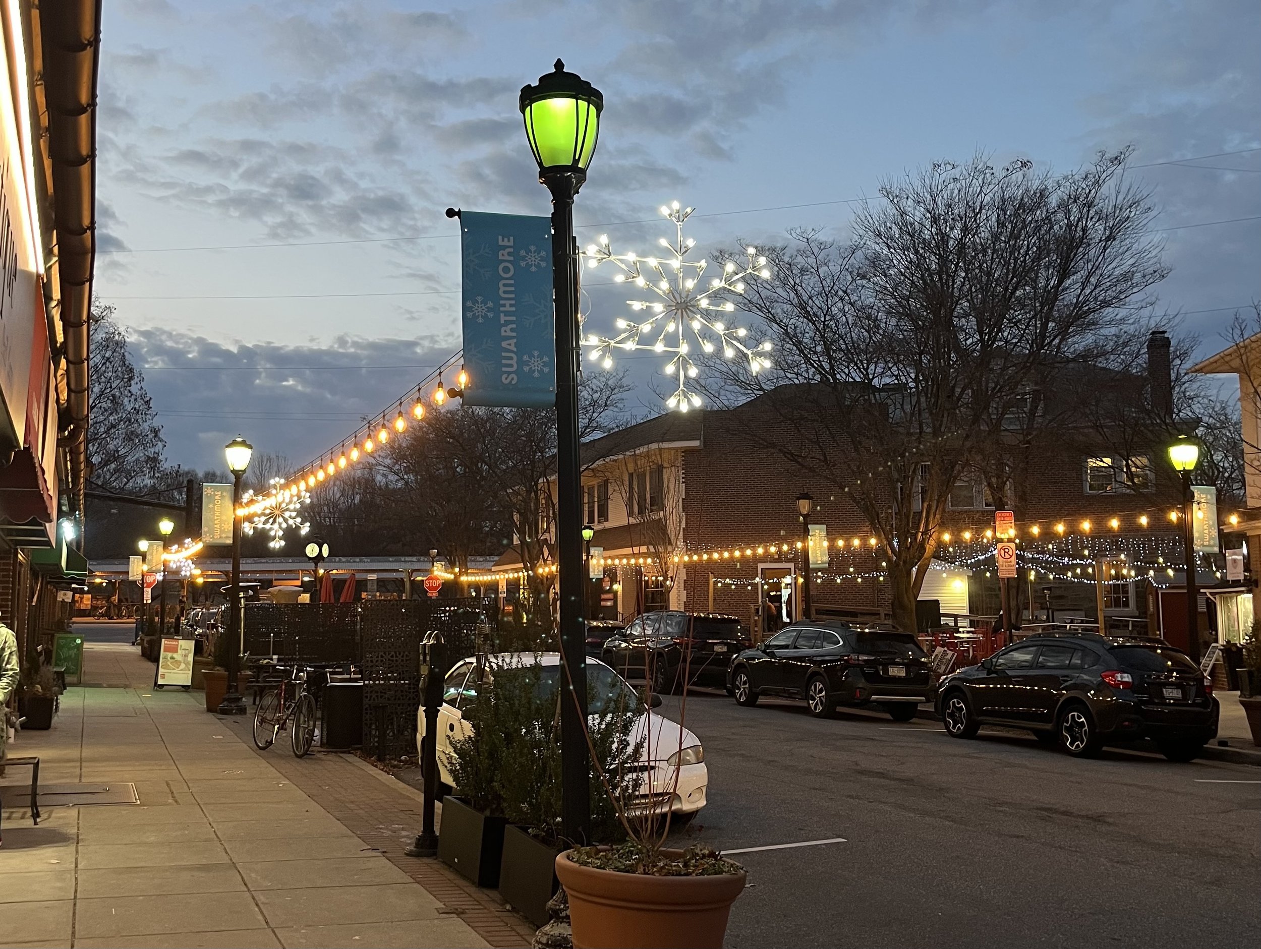 A street with strings of warmly lit light bulbs hung from streetlights along both sides of the street