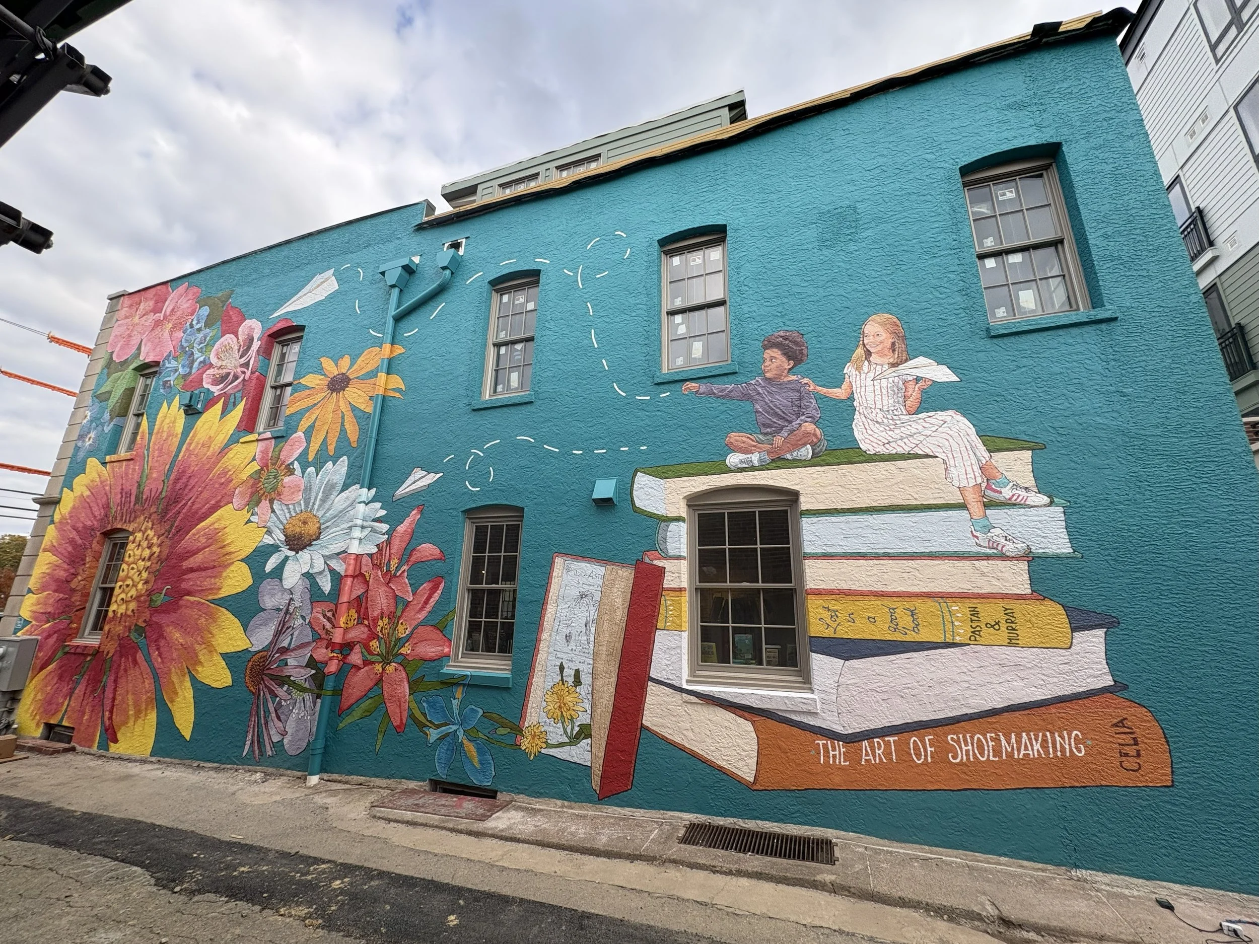 Mural on the side of a building, featuring a blue background with colorful flowers on the left and a larger-than-life stack of books on the right with two children sitting on top.