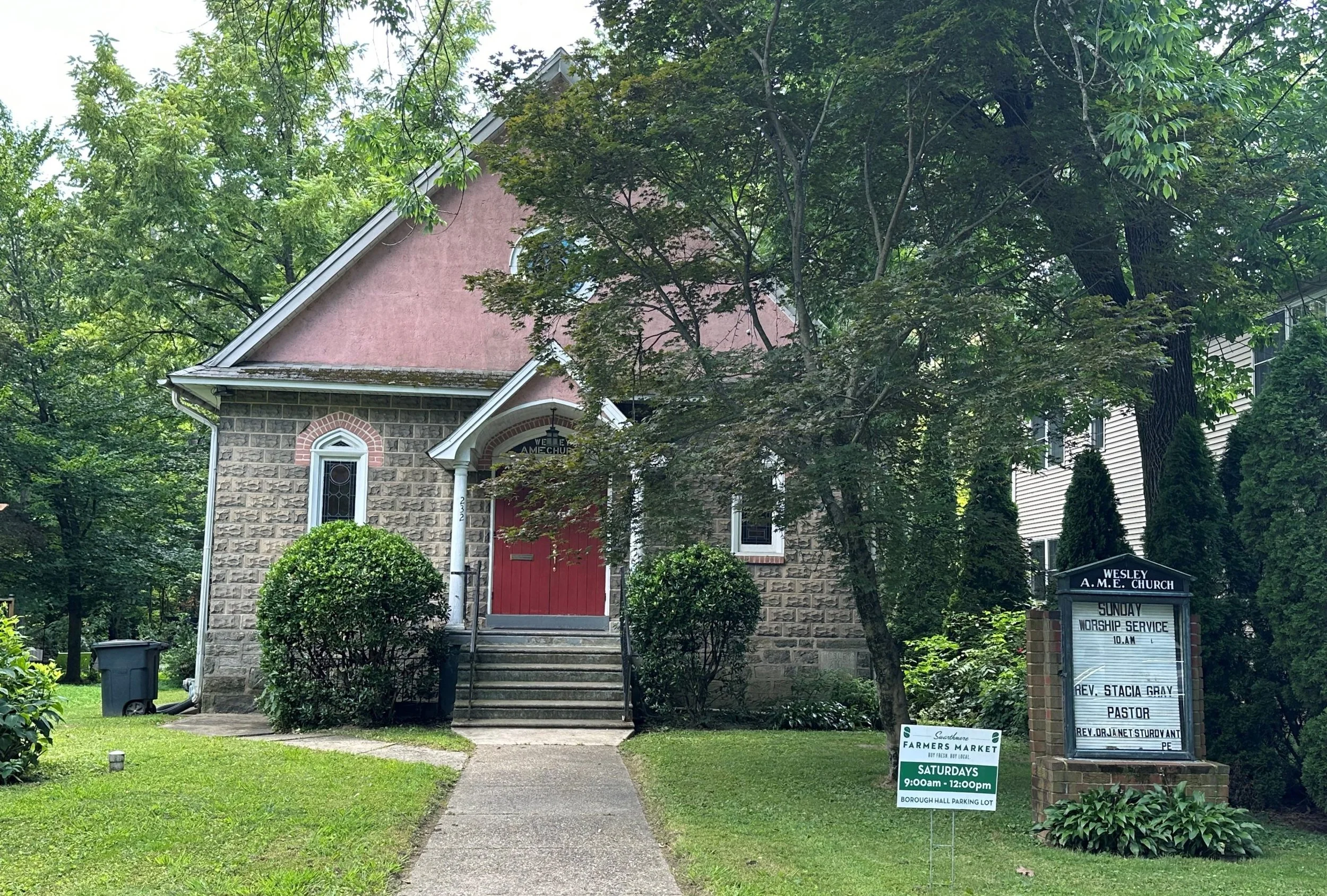 Front facade of a small church building with a pitched roof and steps leading up to a red door. There are trees on the front lawn and a sign welcoming people to Sunday worship service.