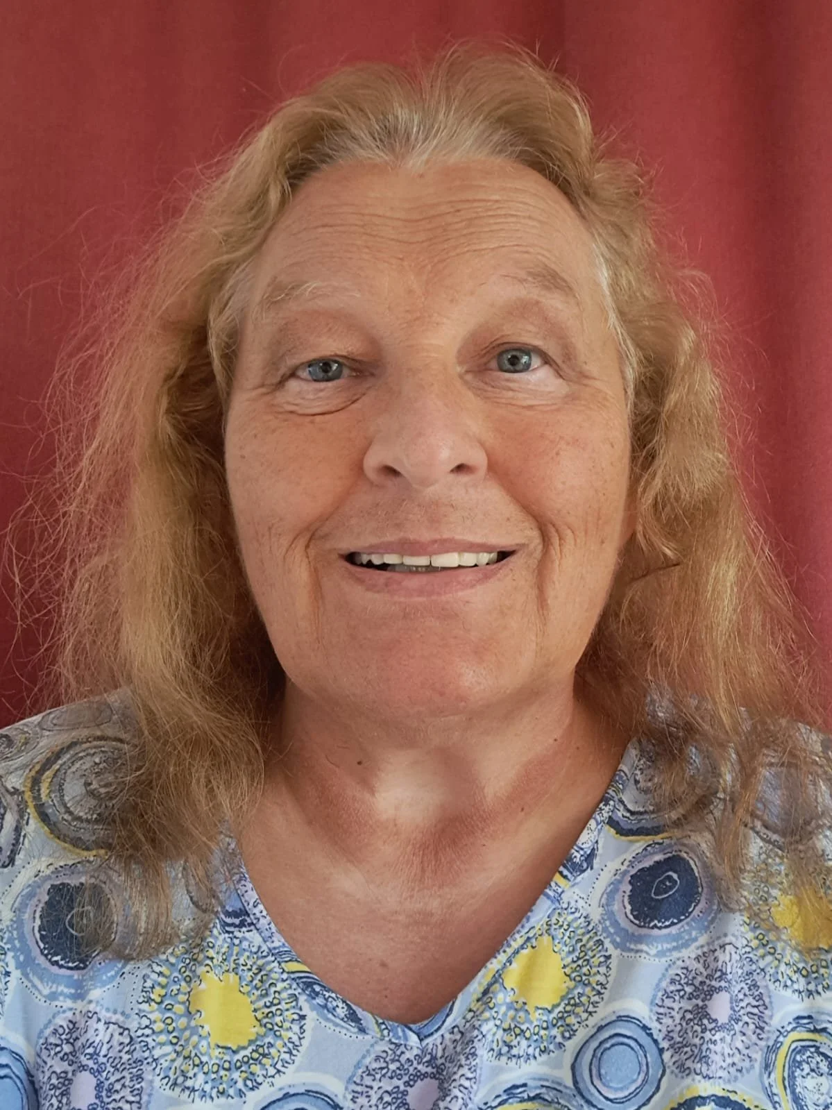 Nahaufnahme einer älteren Frau mit lockigem, blondem Haar vor rotem Hintergrund, trägt ein T-Shirt mit blau-gelben Mustern.