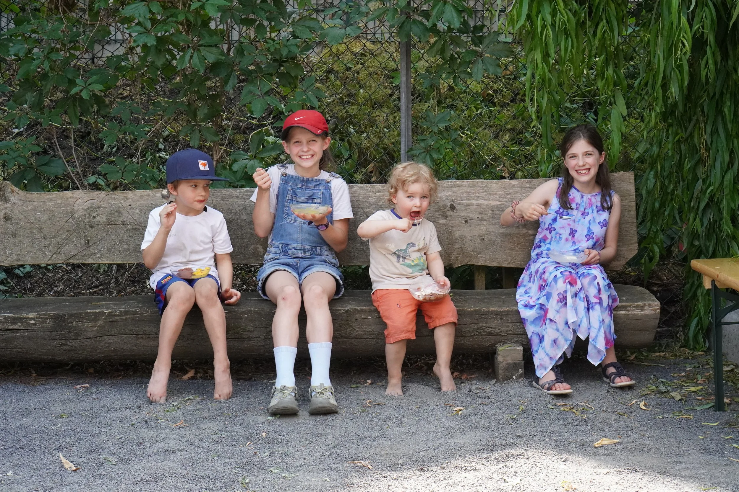 Fünf Kinder sitzen auf einer Holzbank, essen Eis und lachen im Freien, umgeben von Büschen und Bäumen.