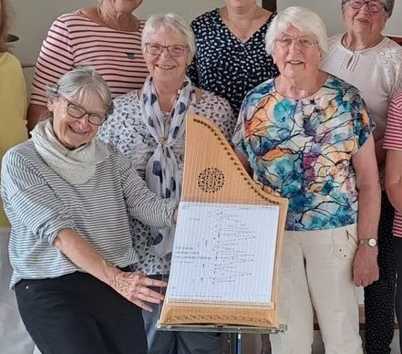 Eine Gruppe älterer Frauen, die zusammen lachen und um ein Cembalo herumstehen, das Notenblätter enthält.