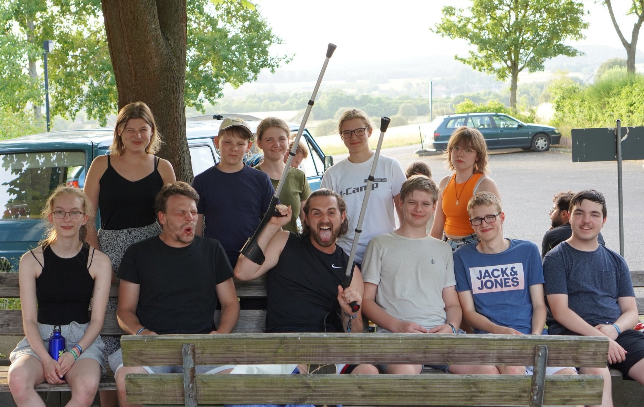 Gruppe junger Leute sitzt auf einer Parkbank im Freien. Einige halten Spazierstöcke, lachen und haben eine fröhliche Stimmung.
