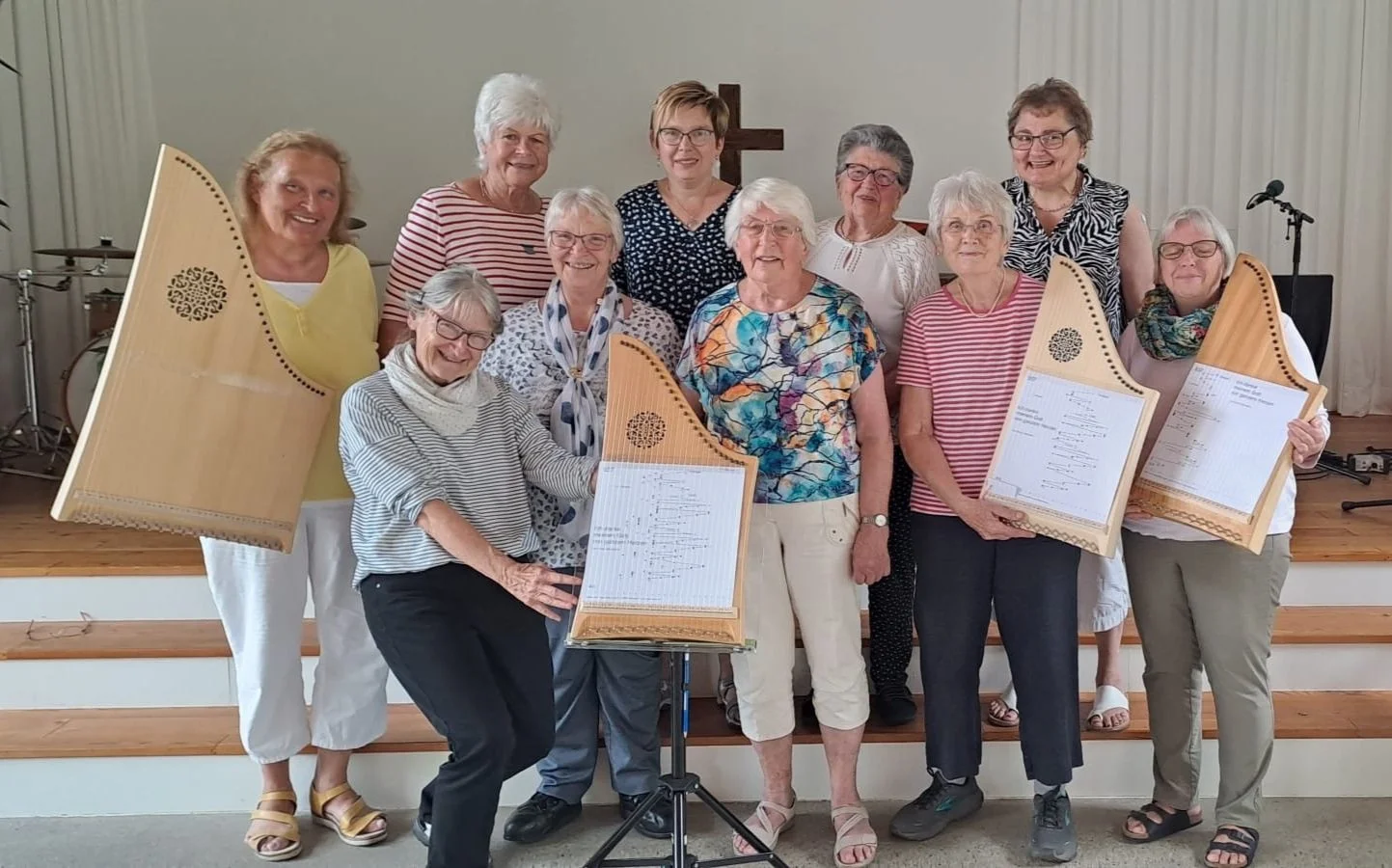 Eine Gruppe älterer Frauen steht zusammen auf einer Bühne, einige halten große Harfen, im Hintergrund ein Kirchenkreuz und Musikinstrumente.