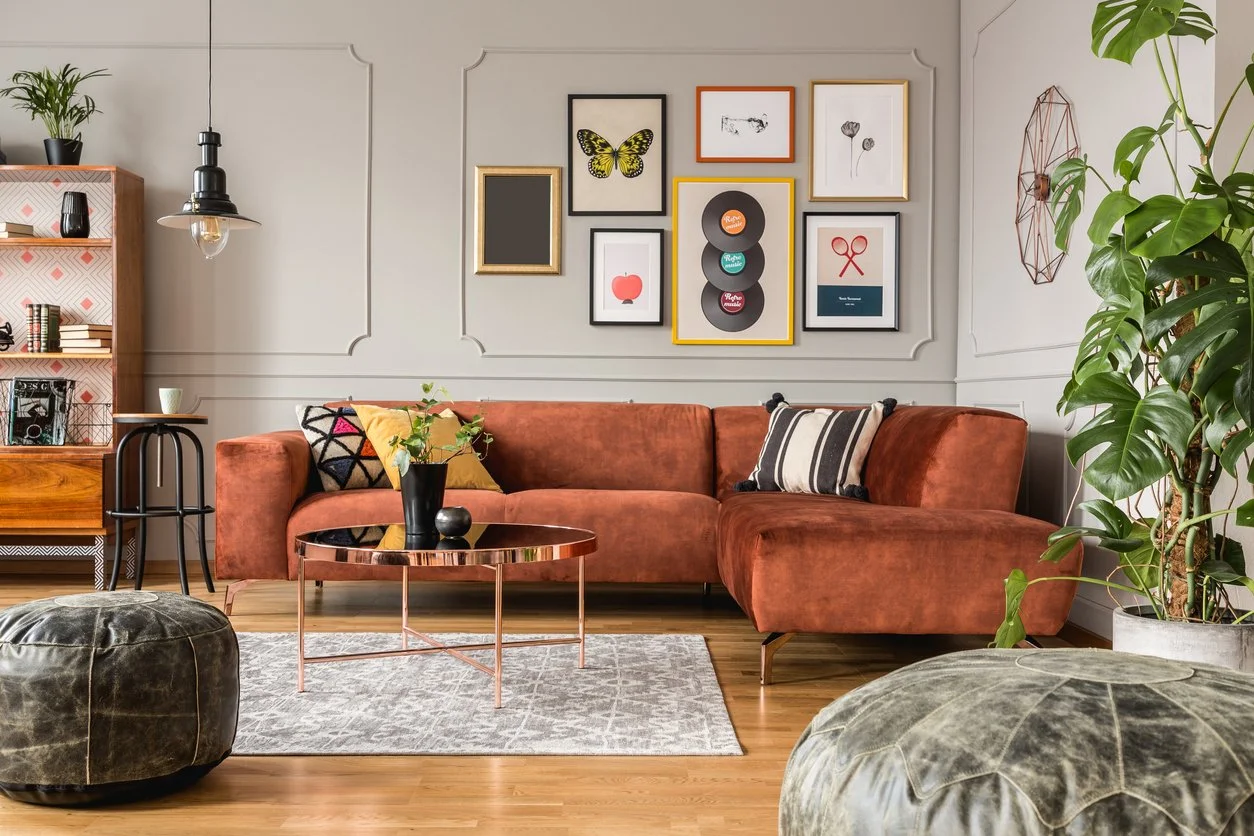 Living room with a salmon-colored velvet sectional sofa, black and striped pillows, and a copper coffee table with a black vase and green plant. A gallery wall with framed art pieces, a large plant on the right, and a wooden bookshelf on the left. Two large poufs in front, and a gray rug on hardwood floors.