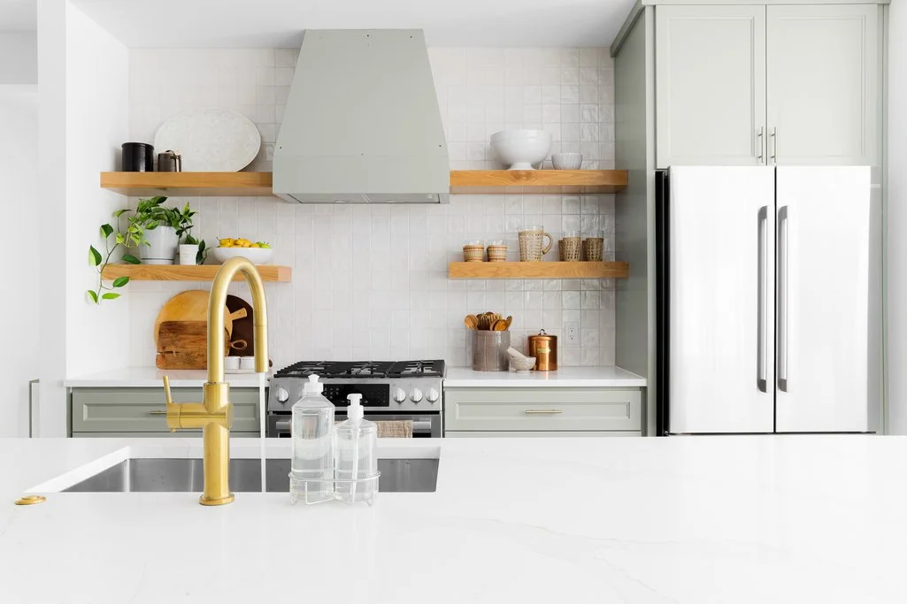 Modern kitchen with light green cabinets, open wooden shelves, white backsplash, gold faucet, and stainless steel appliances.