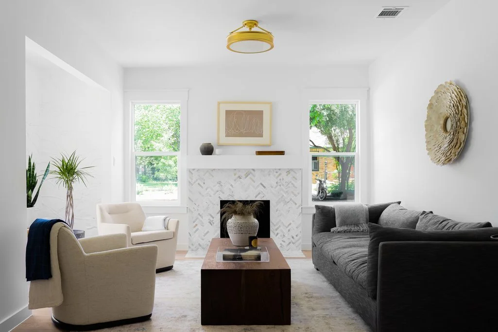 Bright living room with white walls, two large windows, a gray sofa, two beige armchairs, a wooden coffee table, a decorative wall piece, and a fireplace with a white herringbone tile surround.