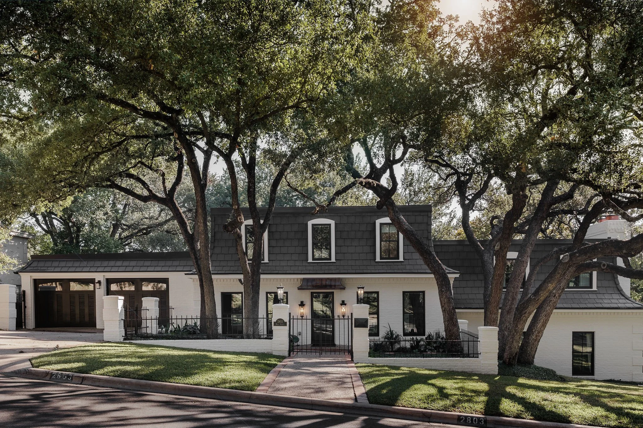 Front exterior of 2803 Wade Ave in Tarrytown, a remodeled 1960s white brick home with dark gray roof, black windows, gated entry, and mature oak trees shading the lawn.