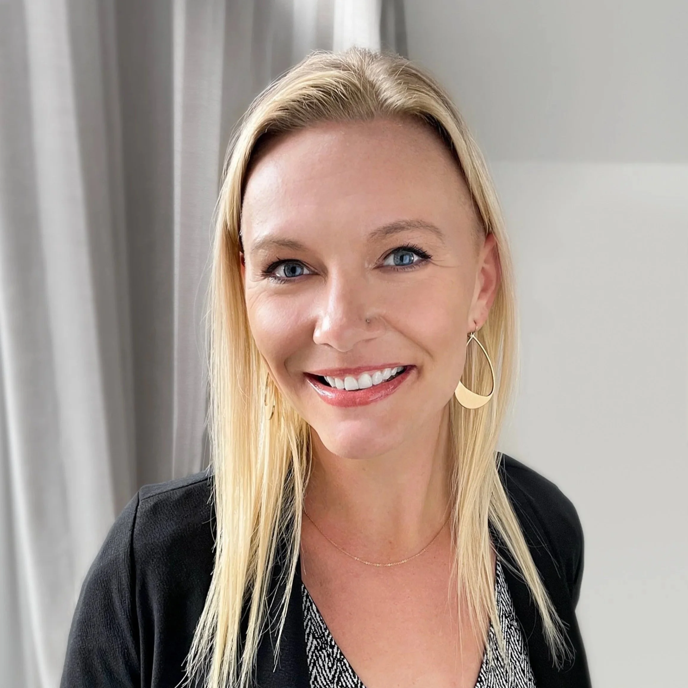 A smiling woman with blonde hair and blue eyes, wearing a black blazer, patterned blouse, gold hoop earrings, and a delicate necklace, standing in front of light-colored curtains.