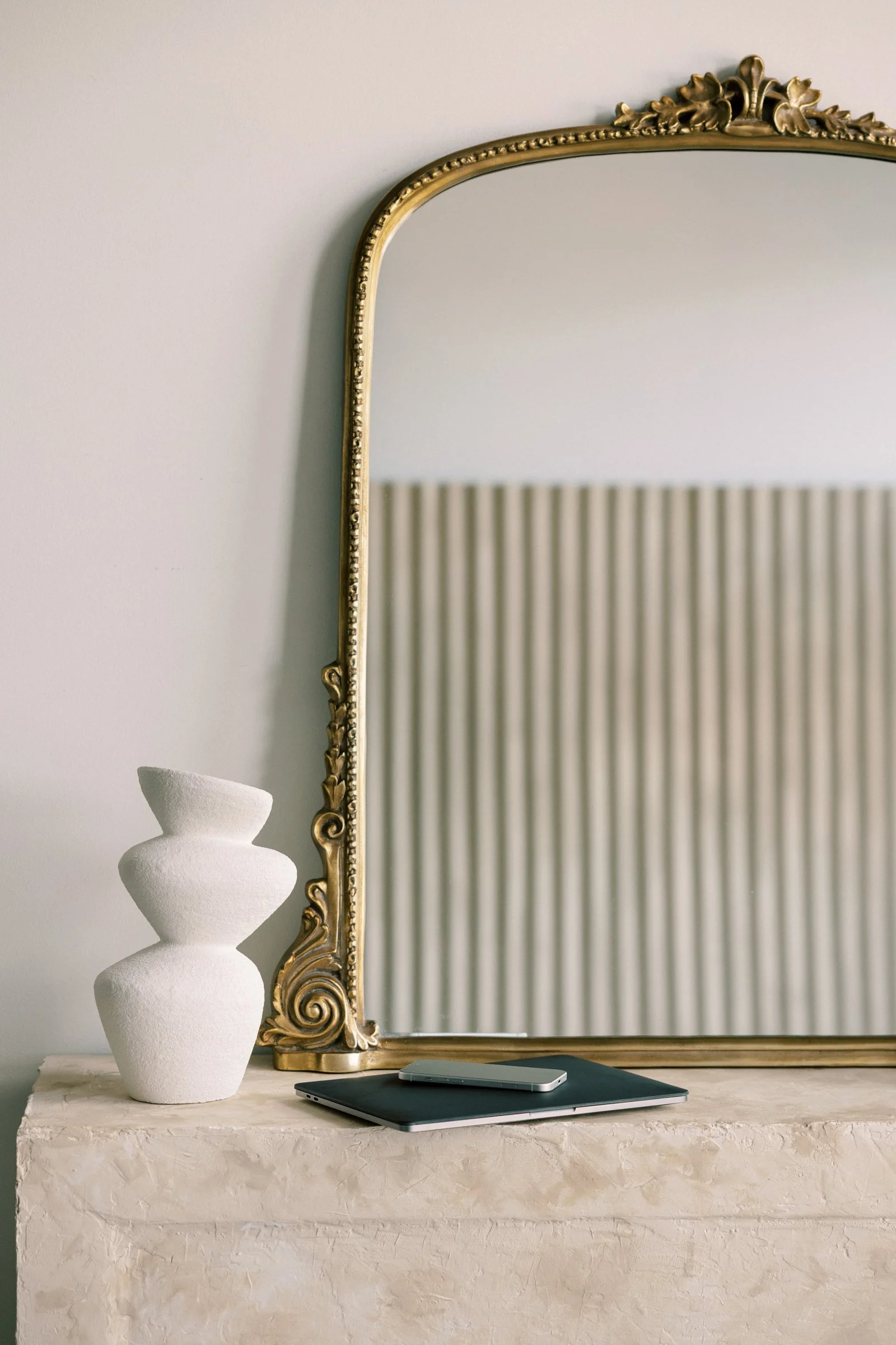 Decorative gold-framed mirror on a wall, a white textured vase with three stacked rounded shapes, a closed dark green laptop, and a closed silver tablet or smartphone on a light-colored textured stone surface.
