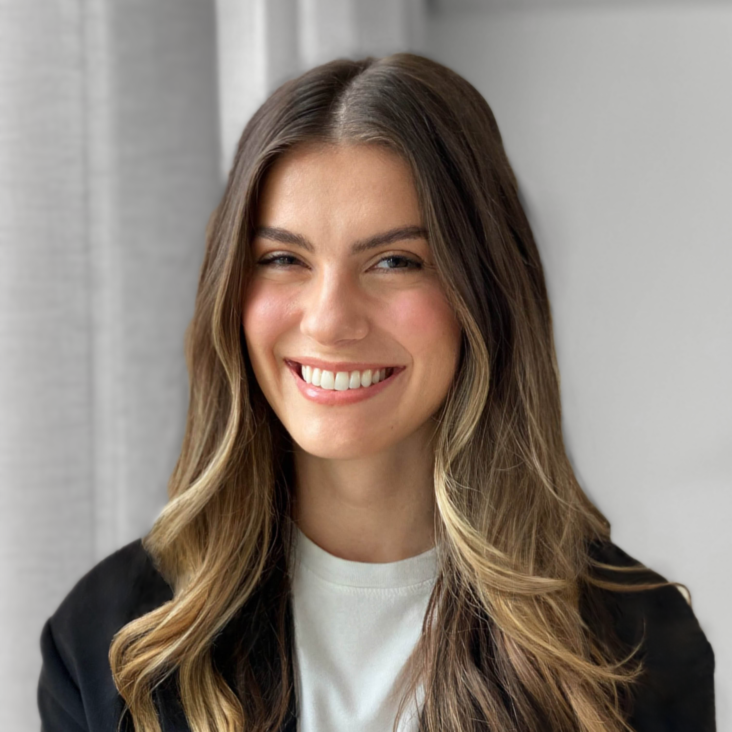 A woman with long wavy brown hair smiling, wearing a white shirt and black blazer, standing near a light-colored wall.