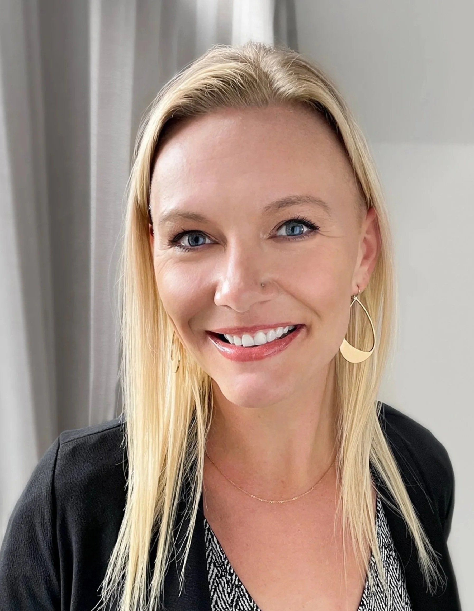 A smiling woman with long blonde hair and blue eyes, wearing large hoop earrings, a black jacket, and a patterned top, standing indoors near gray curtains.