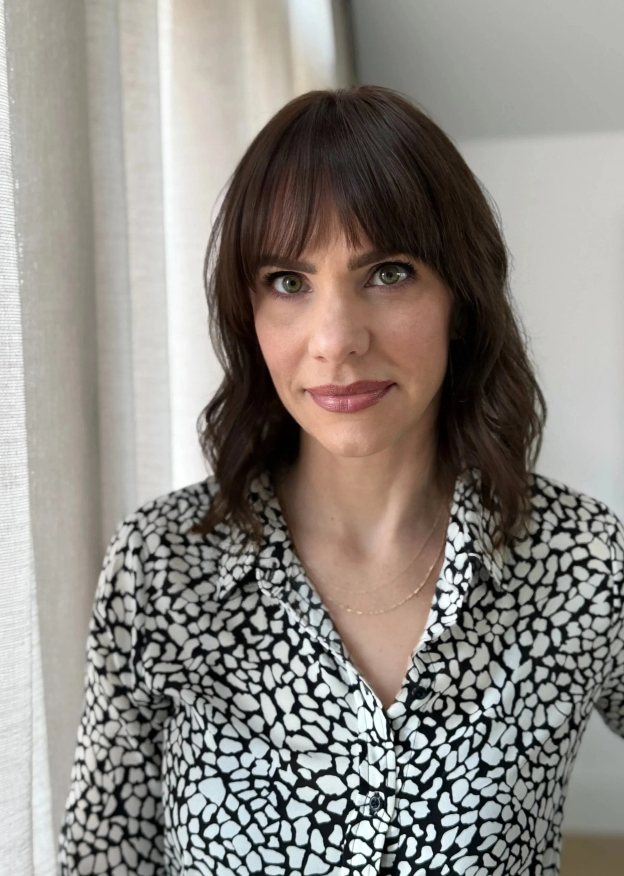 A woman with shoulder-length brown hair and green eyes wearing a black and white patterned blouse, standing indoors next to a window with light-colored curtains.