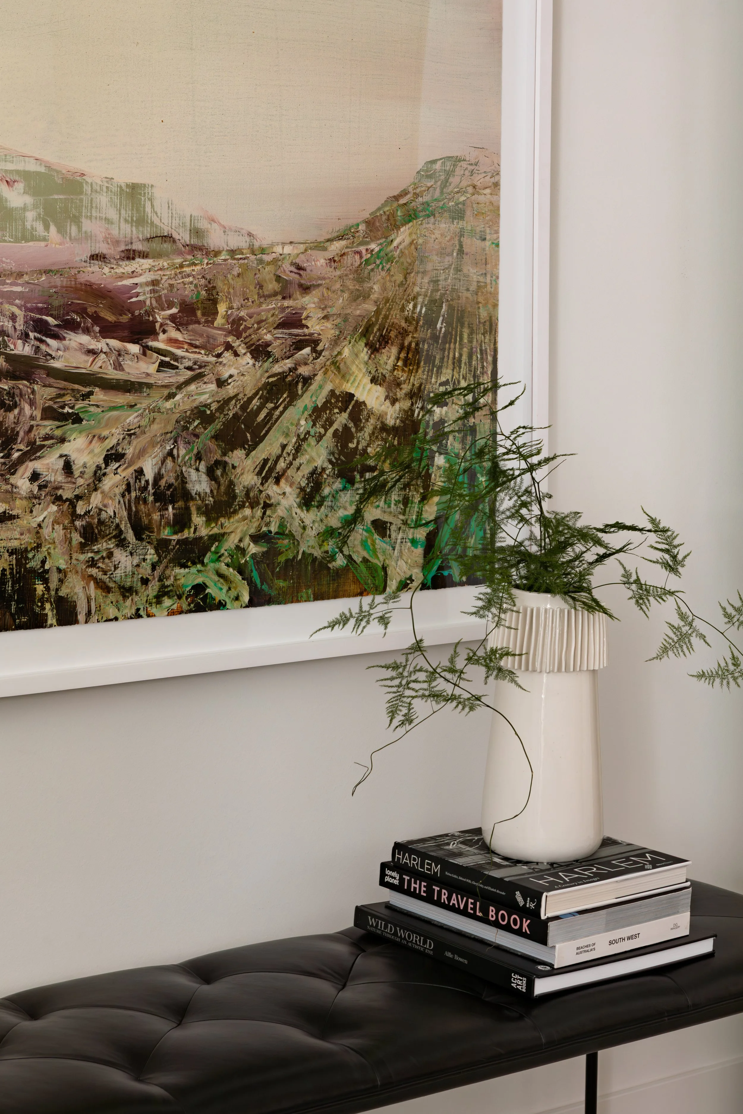 A black leather tufted bench with a stack of four travel books on top. A white vase with a green leafy plant is placed on the books. A large abstract landscape painting hangs on a white wall behind the bench.