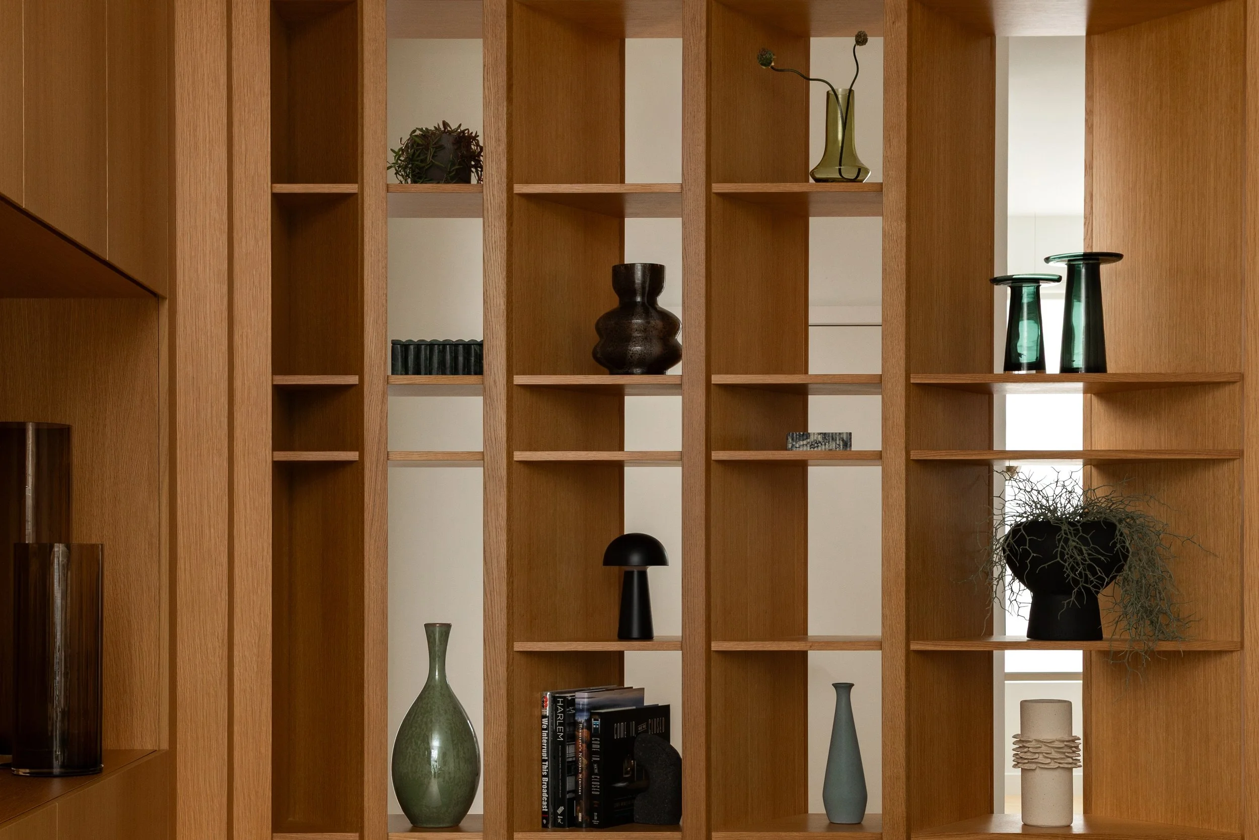 A wooden room divider with shelves holds various decorative vases and plants, with some books and a black sculpture at the bottom.