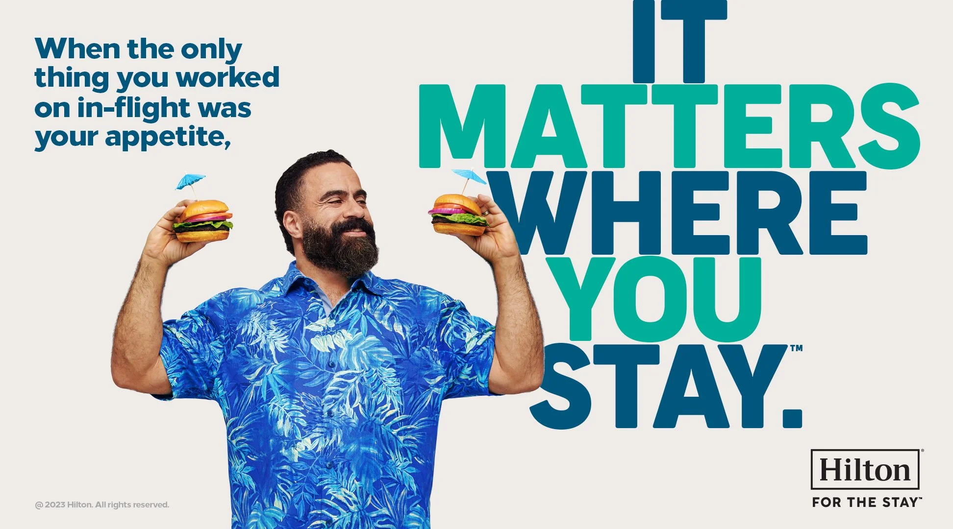 A man wearing a blue floral shirt holding two small hamburgers with umbrellas above them, smiling with eyes closed, with a text overlay about the importance of staying in place when working on in-flight meals, and a Hilton logo at the bottom right corner.