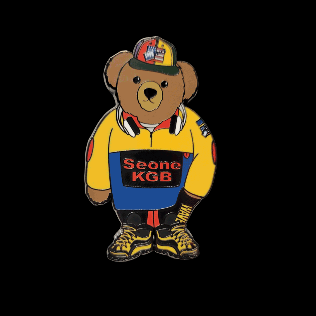 A teddy bear wearing a yellow jacket labeled "Seone KGB," black pants, a colorful cap, headphones around its neck, and yellow shoes. The background is black.