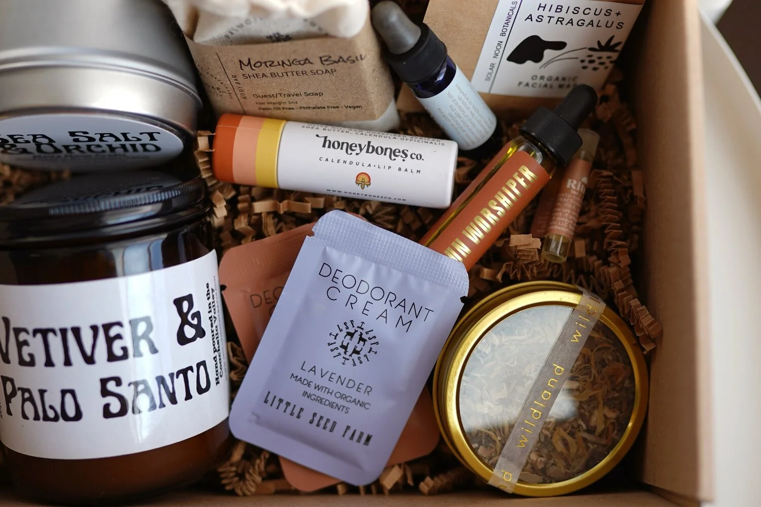 A gift box containing various natural skincare, wellness, and aromatherapy products, including a jar labeled 'Netiver & Palo Santo,' a lavender deodorant cream packet, a mini face roller, a tincture bottle, a lip balm, a spray bottle, and a container