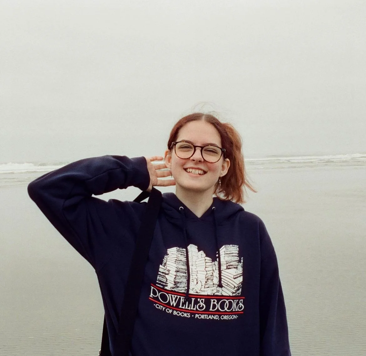 A young woman with red hair and glasses smiling while standing on a beach with the ocean in the background, wearing a navy hoodie with 'Powell's Books' logo.