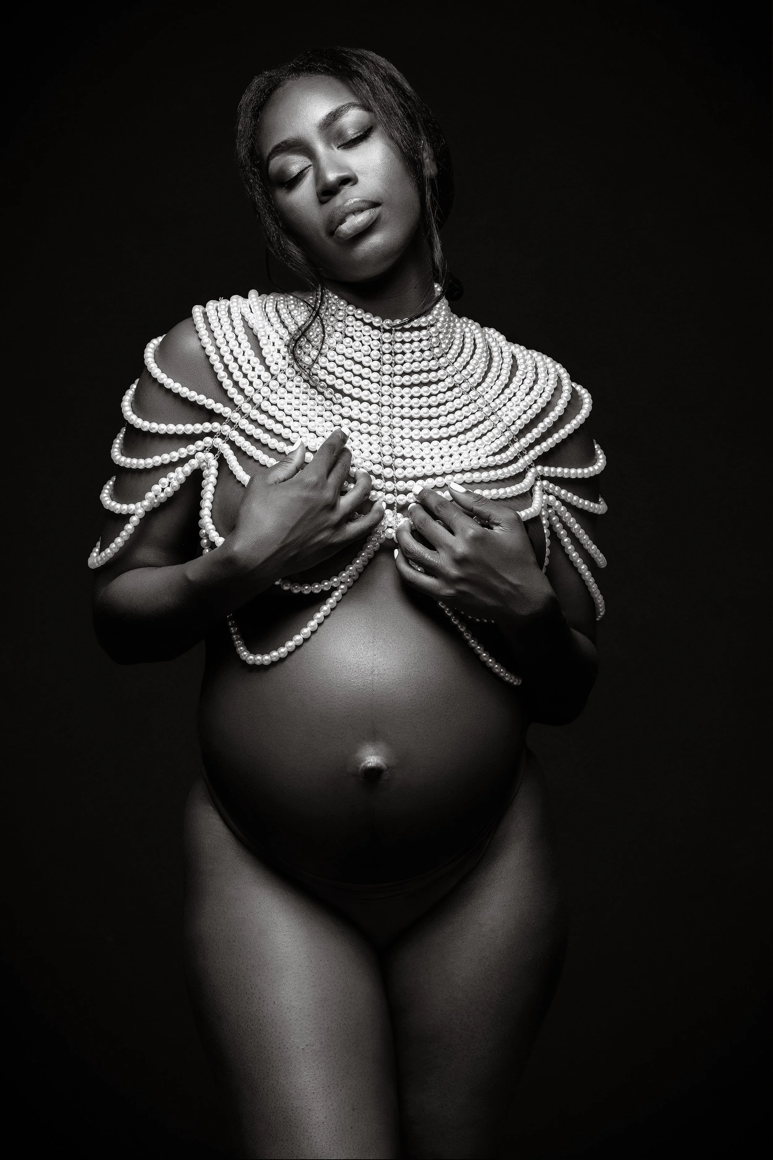 Black and white portrait of an elegant woman with jewelry, her eyes closed and hands on her chest, against a dark background.