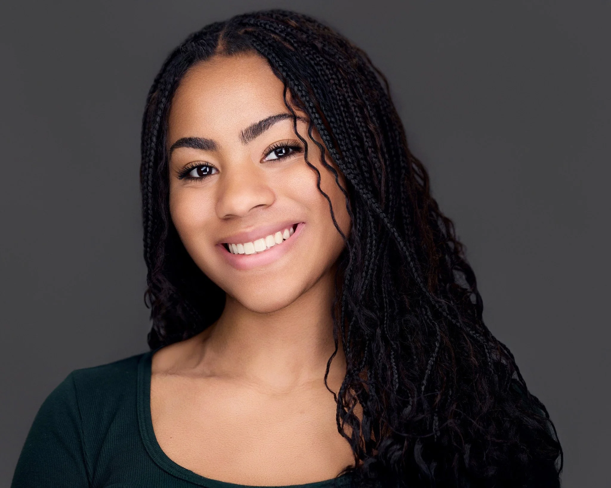 A young woman smiling with long curly hair wearing a dark green top against a gray background.