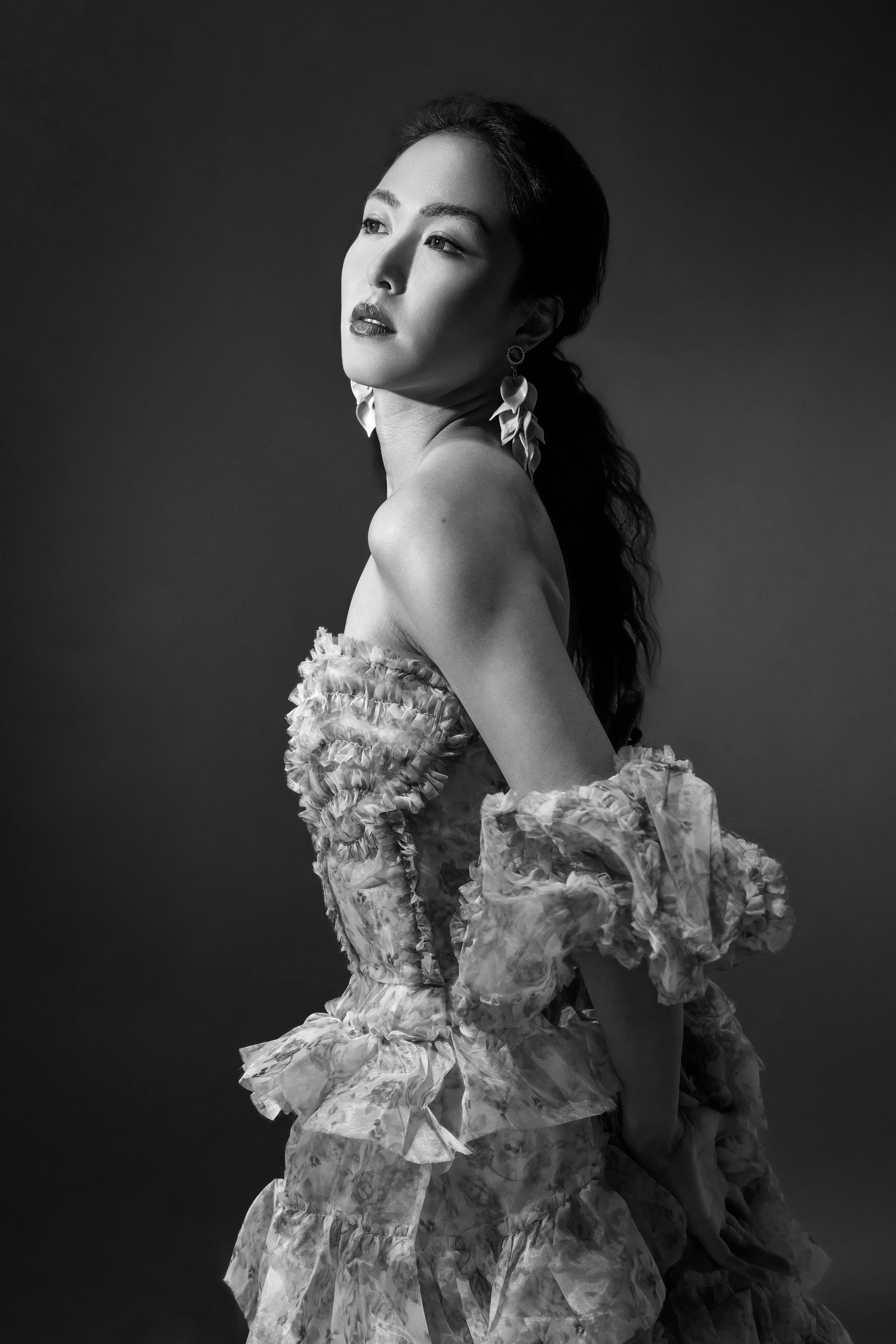 Black and white portrait of a woman with long wavy hair, wearing statement earrings and a ruffled, textured dress with puffy sleeves, posing with her shoulders angled and looking slightly upward.